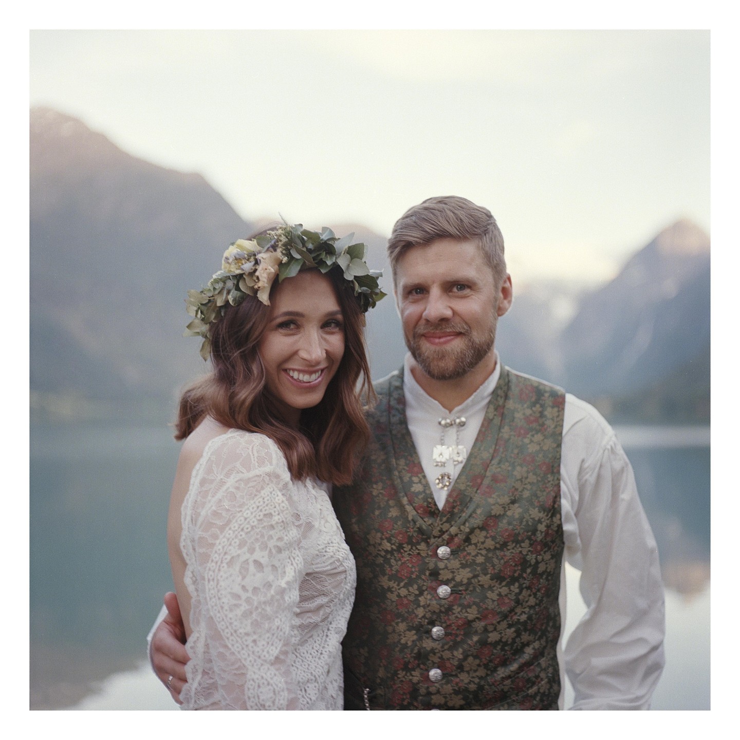 A couple stands by a lake with mountains in the background.