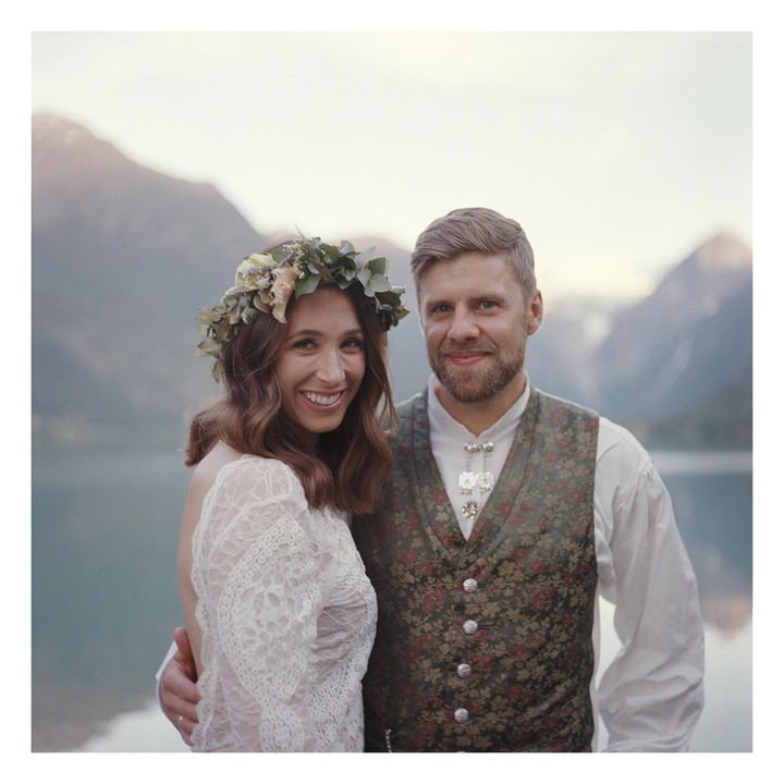 A couple stands by a lake with mountains in the background.