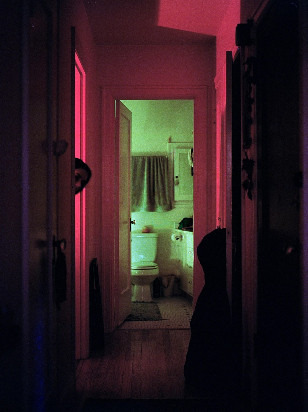 A person peeks from behind a door in a hallway with a bathroom visible at the end.