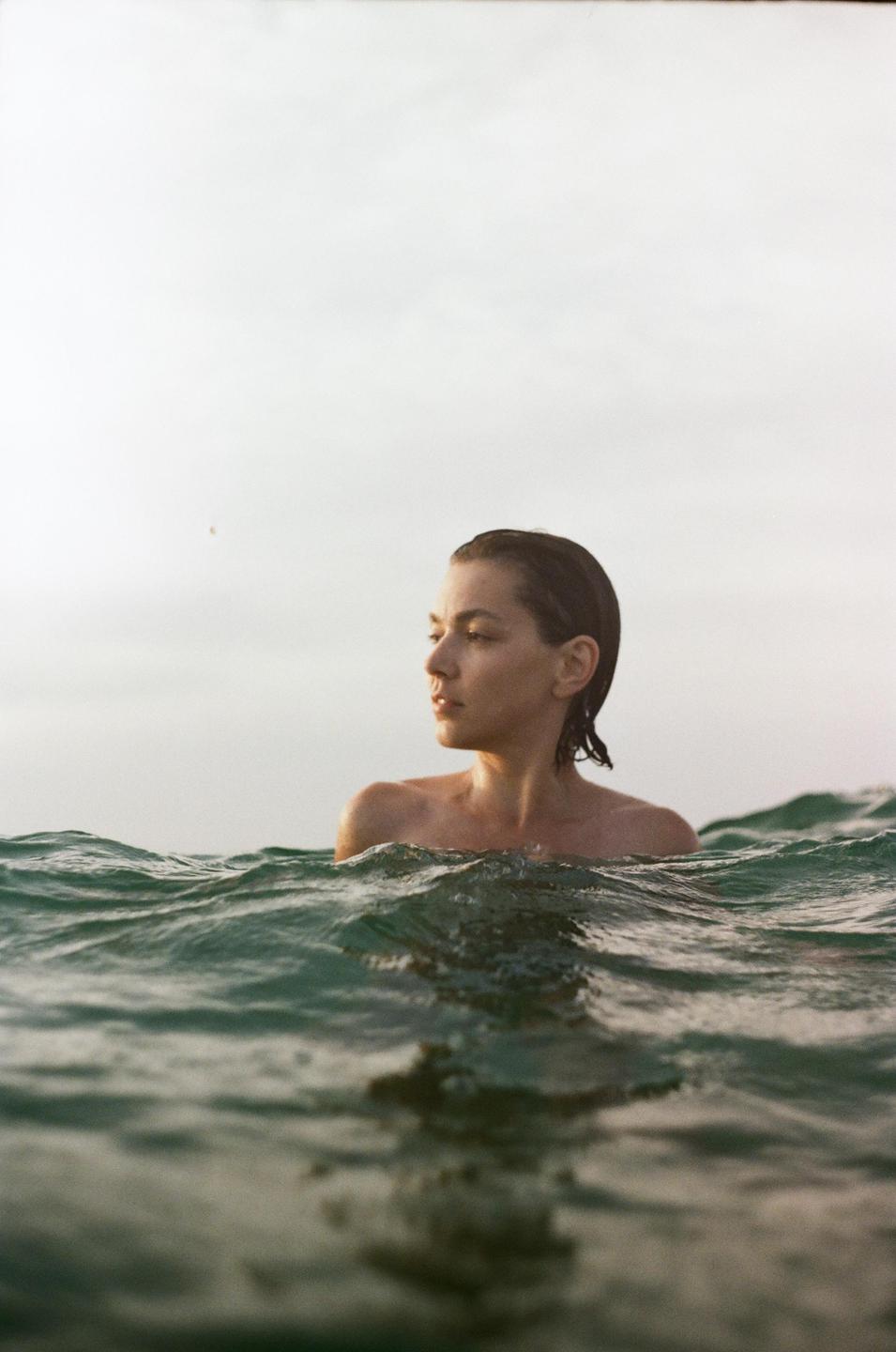 A woman with wet hair is in the ocean with waves around her shoulders.