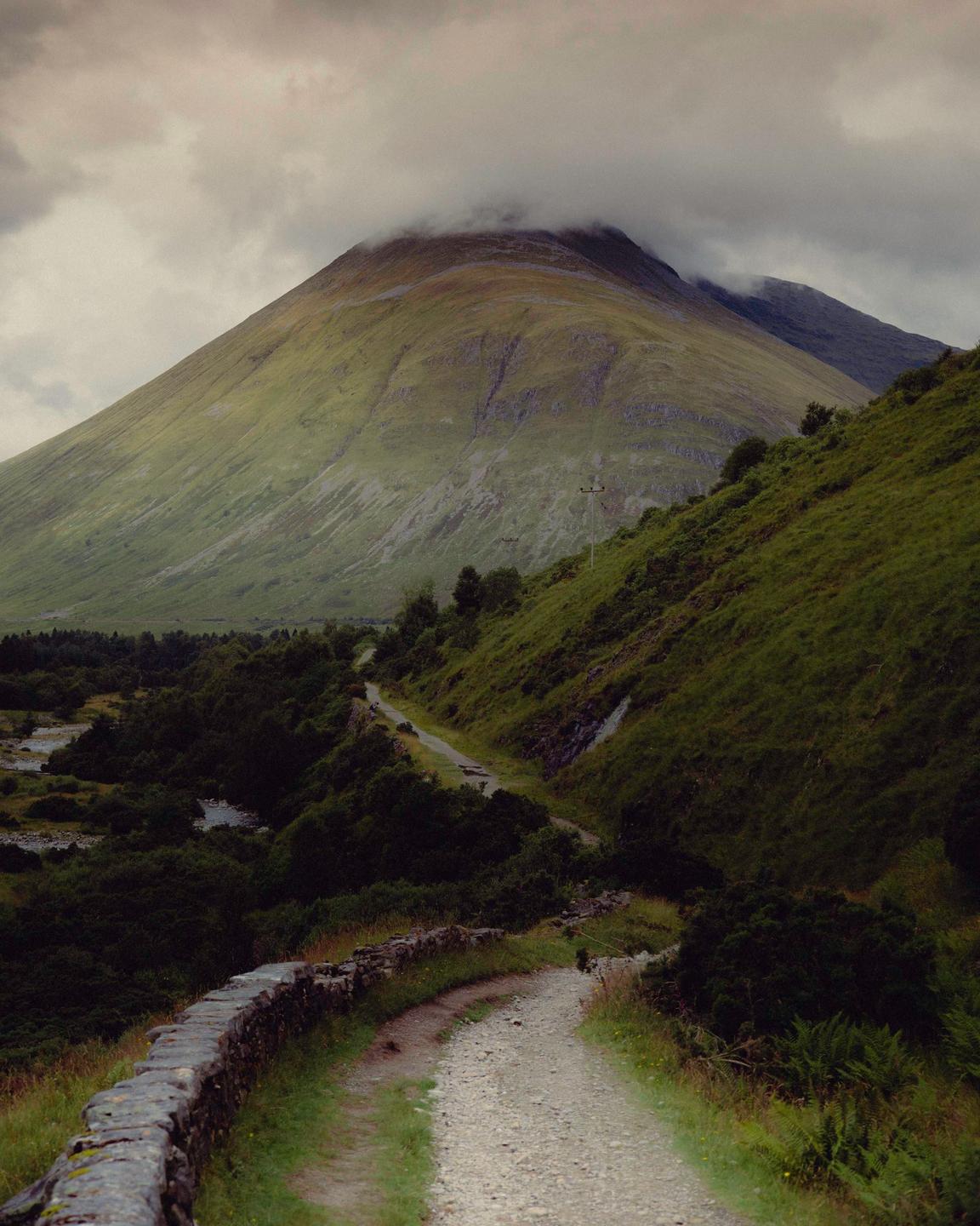A winding path leads through a valley towards a large mountain partially obscured by clouds.