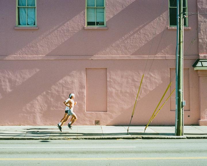Two men jog along a sidewalk next to a pink building with windows and a utility pole.