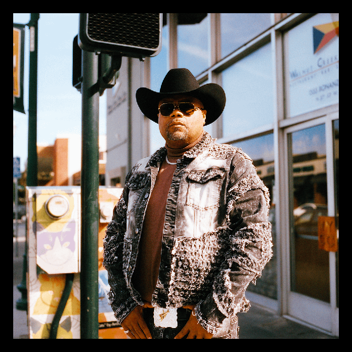 A man wearing a cowboy hat and sunglasses stands on a street in front of a building.