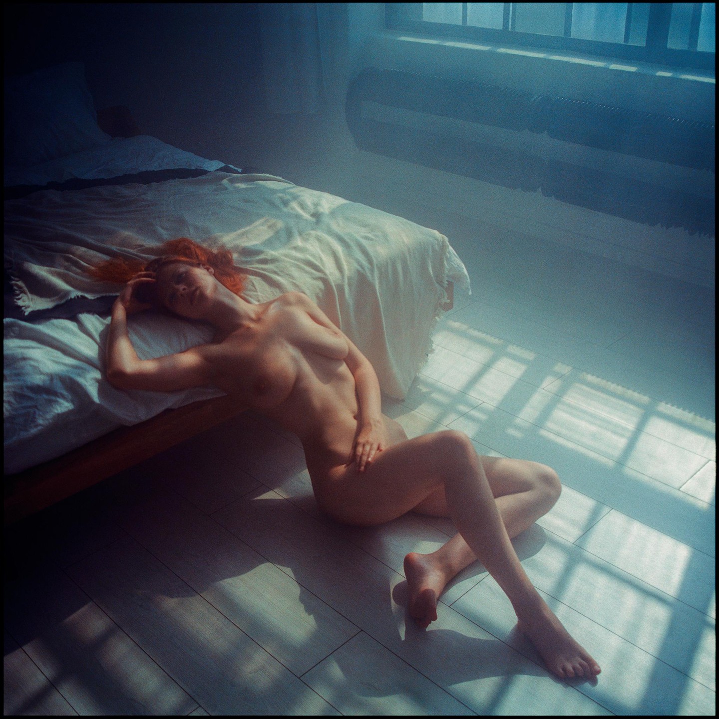 A nude woman with red hair is lying on the floor next to a bed, illuminated by window light.