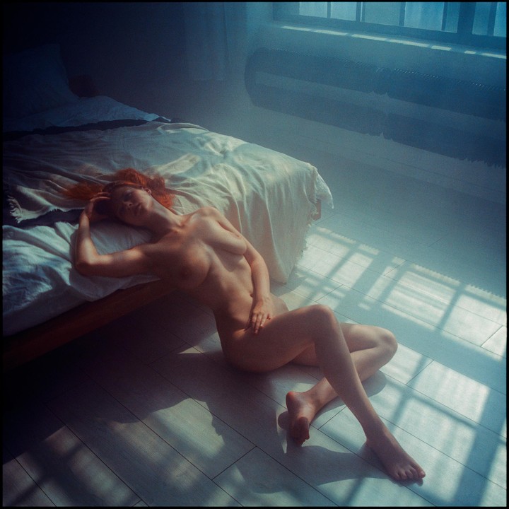 A nude woman with red hair is lying on the floor next to a bed, illuminated by window light.