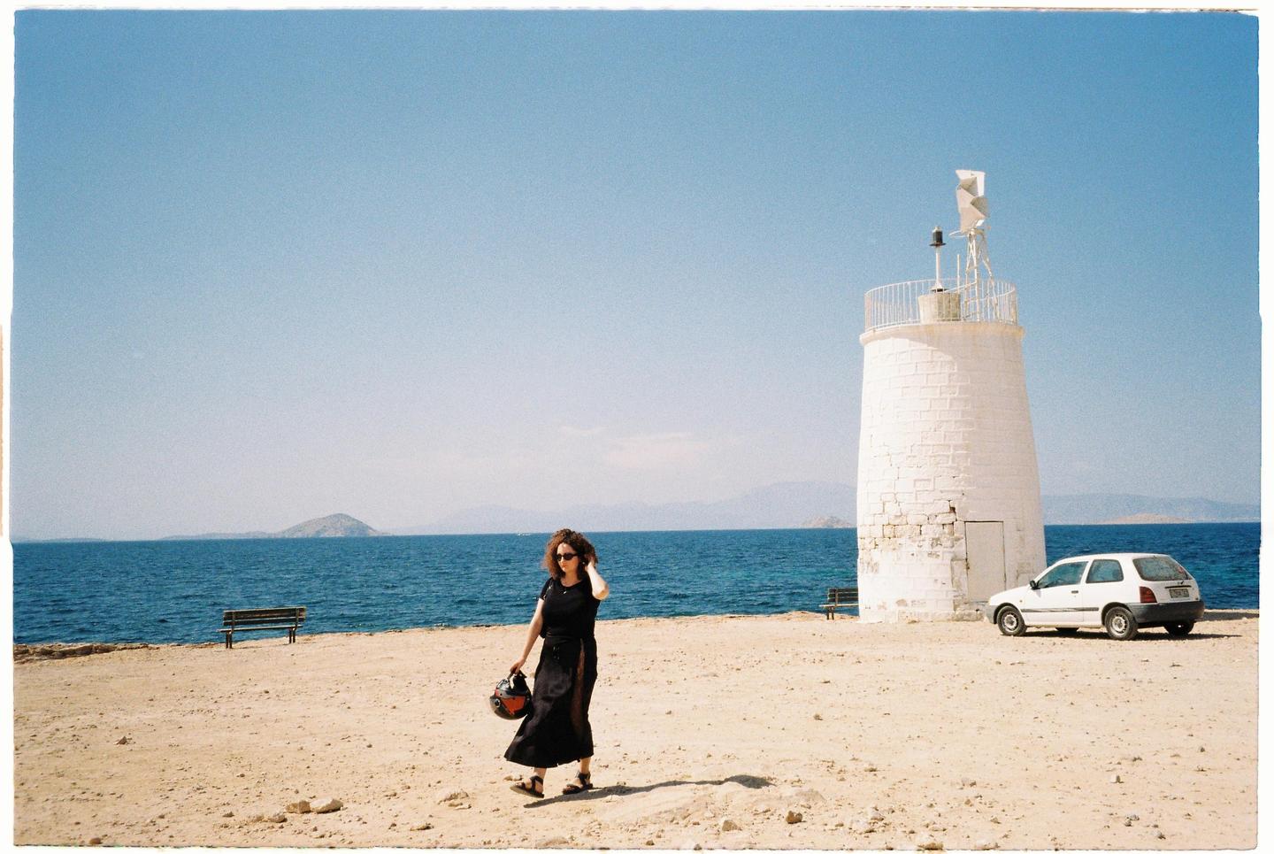 A woman walks on a sandy beach next to a white lighthouse and the sea.