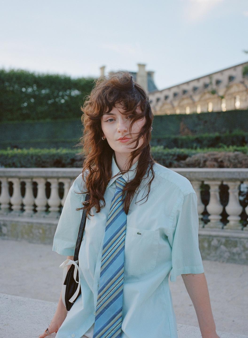 A woman with long, curly hair and a striped tie poses outdoors with a balustrade and building in the background.