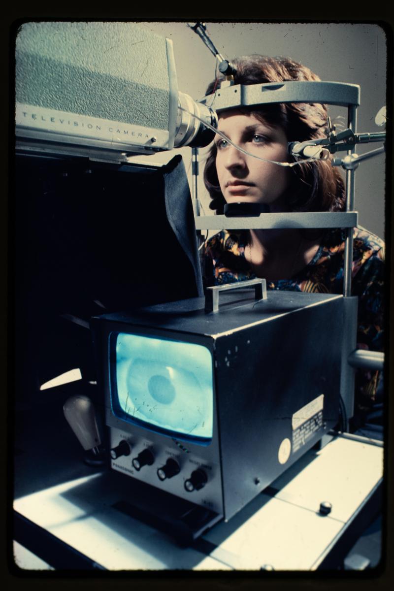A woman is looking into an eye-tracking device with a television camera and a monitor displaying an eye.