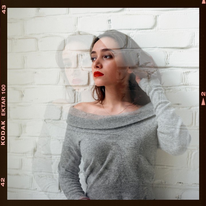 A woman with red lipstick and a gray sweater is captured in a double exposure against a white brick wall.