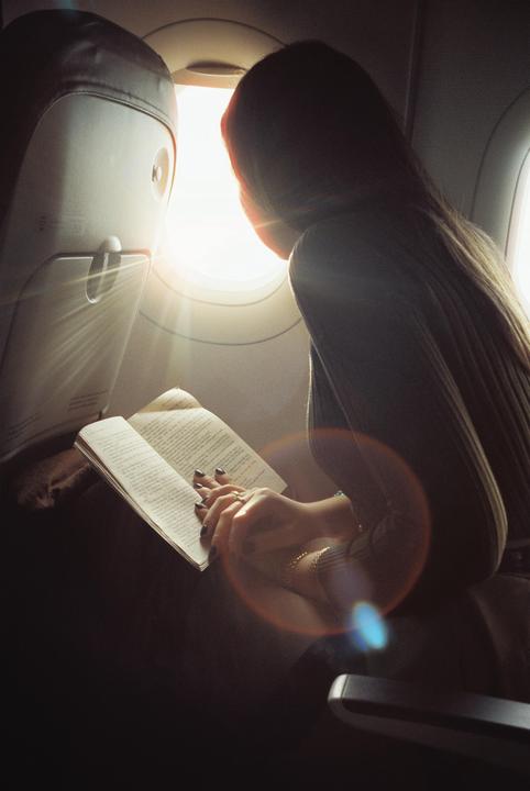 A woman reads a book while sitting in an airplane seat next to a window with bright light coming through.