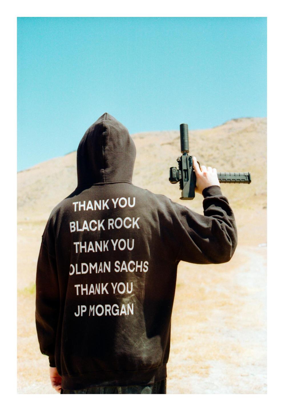 A person wearing a black hoodie with white text on the back holds a firearm in a desert landscape.