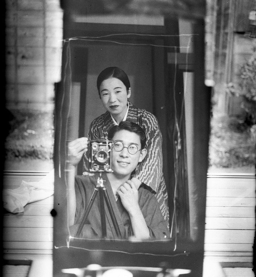 A man in glasses holds a camera on a tripod while a woman stands behind him, both reflected in a mirror.