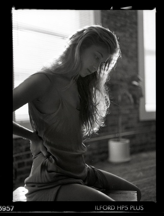 A woman with long hair sits in profile, looking down, with light coming from a window behind her.