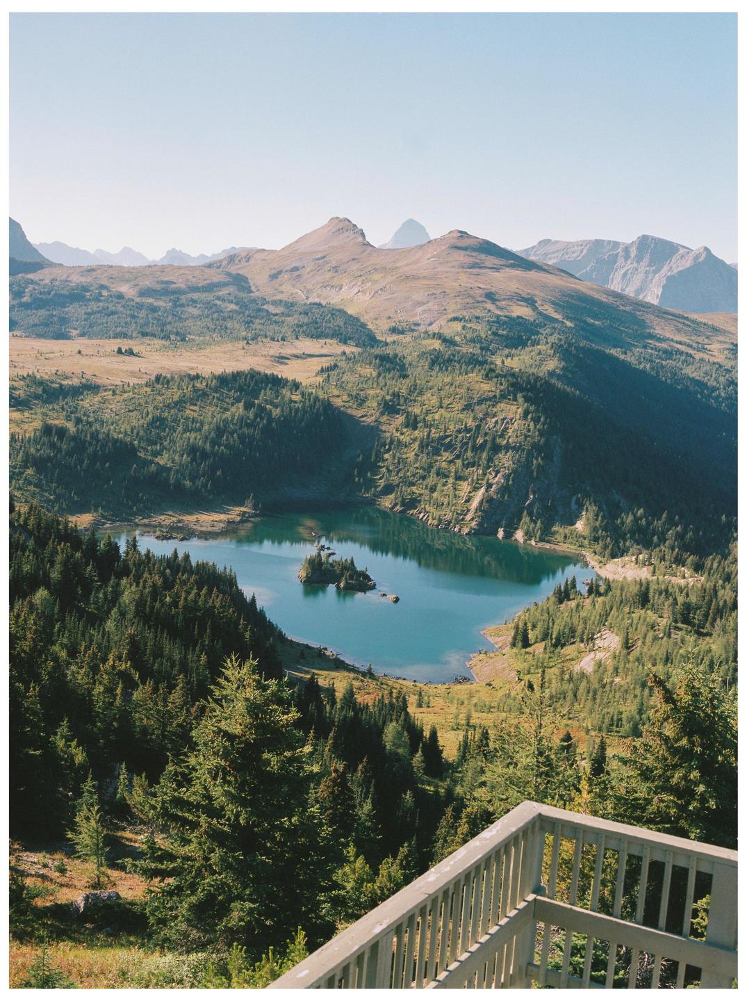 A mountain lake with a small island is surrounded by a forest and mountains under a clear sky.