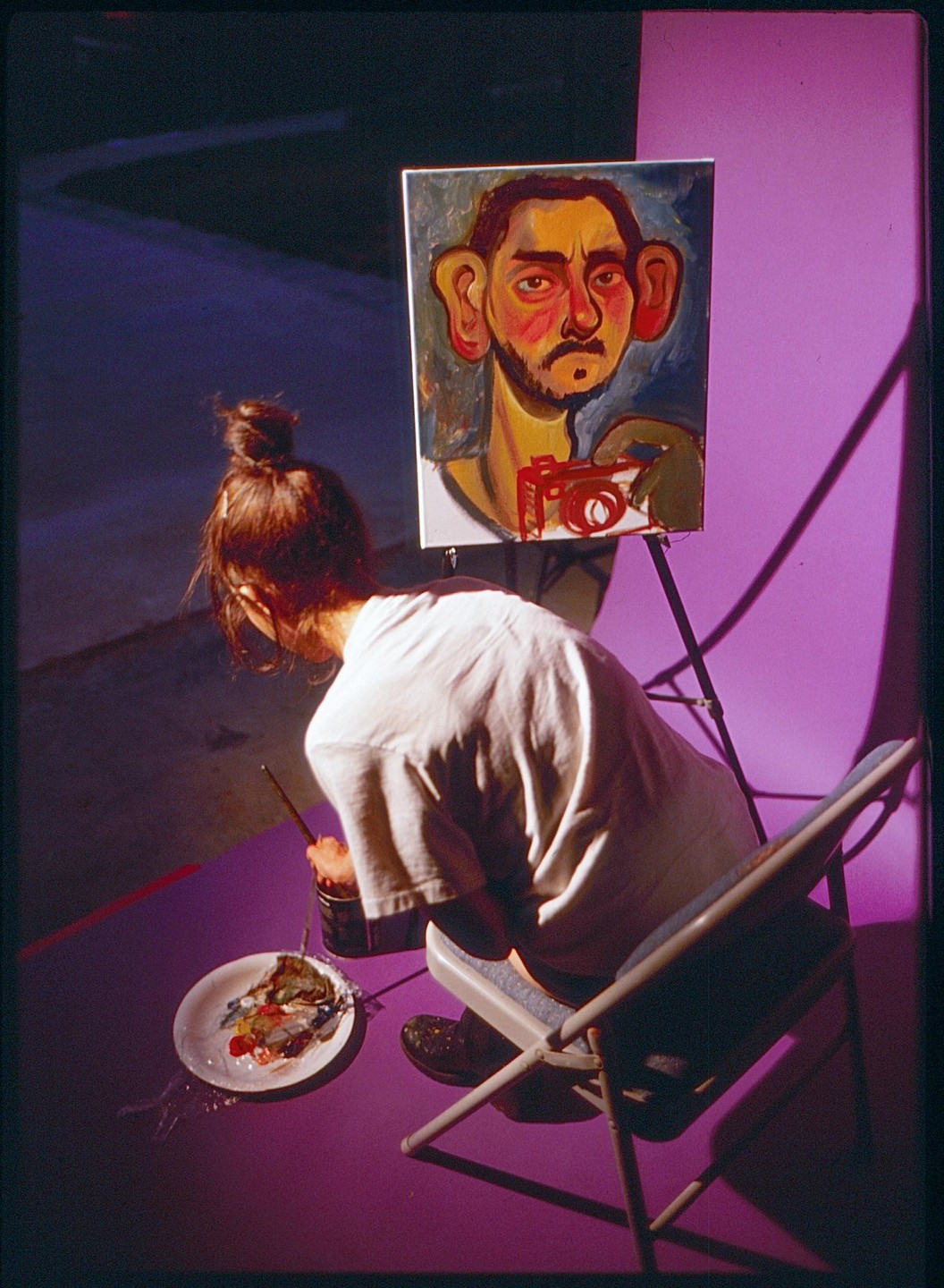 A woman sits on a folding chair painting a self-portrait on an easel.