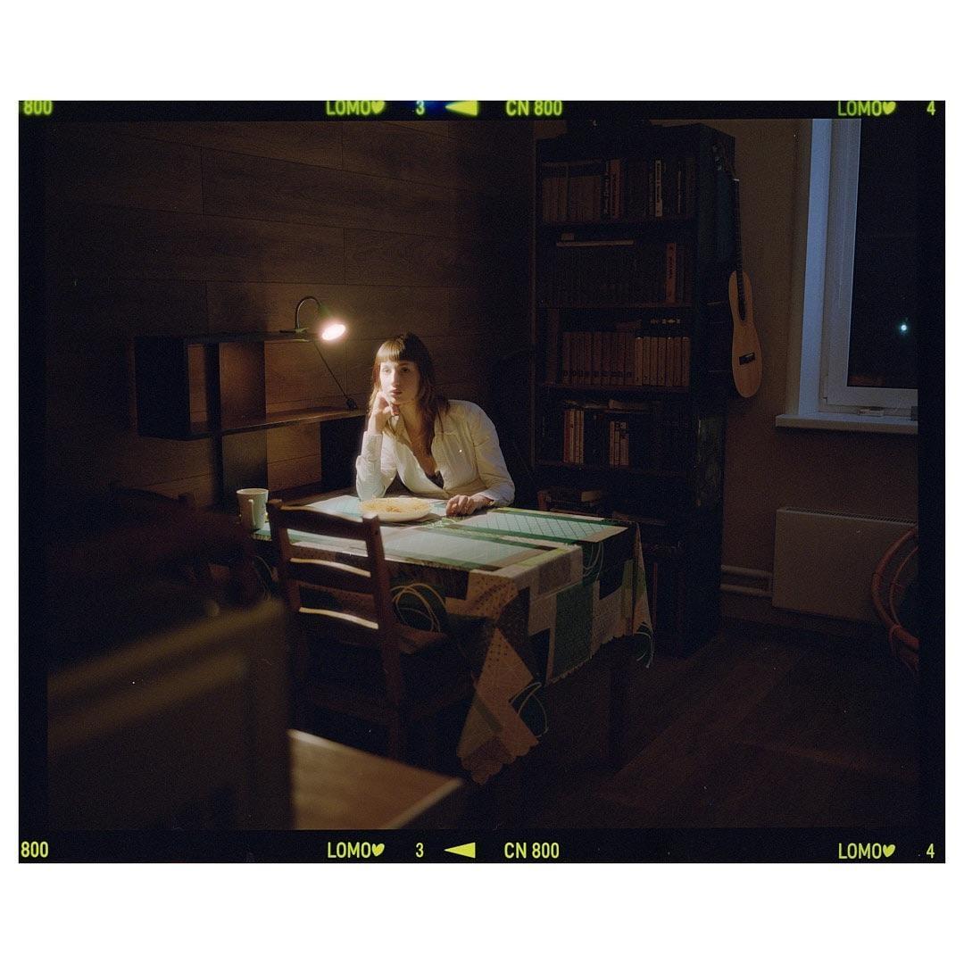 A woman sits at a table with a plate of food, illuminated by a desk lamp, in a dimly lit room.