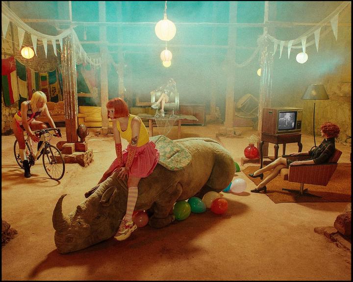 Three women are in a hazy room with a rhinoceros, balloons, and a television.