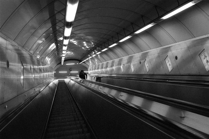 A person sits on an escalator in a long, brightly lit underground tunnel.