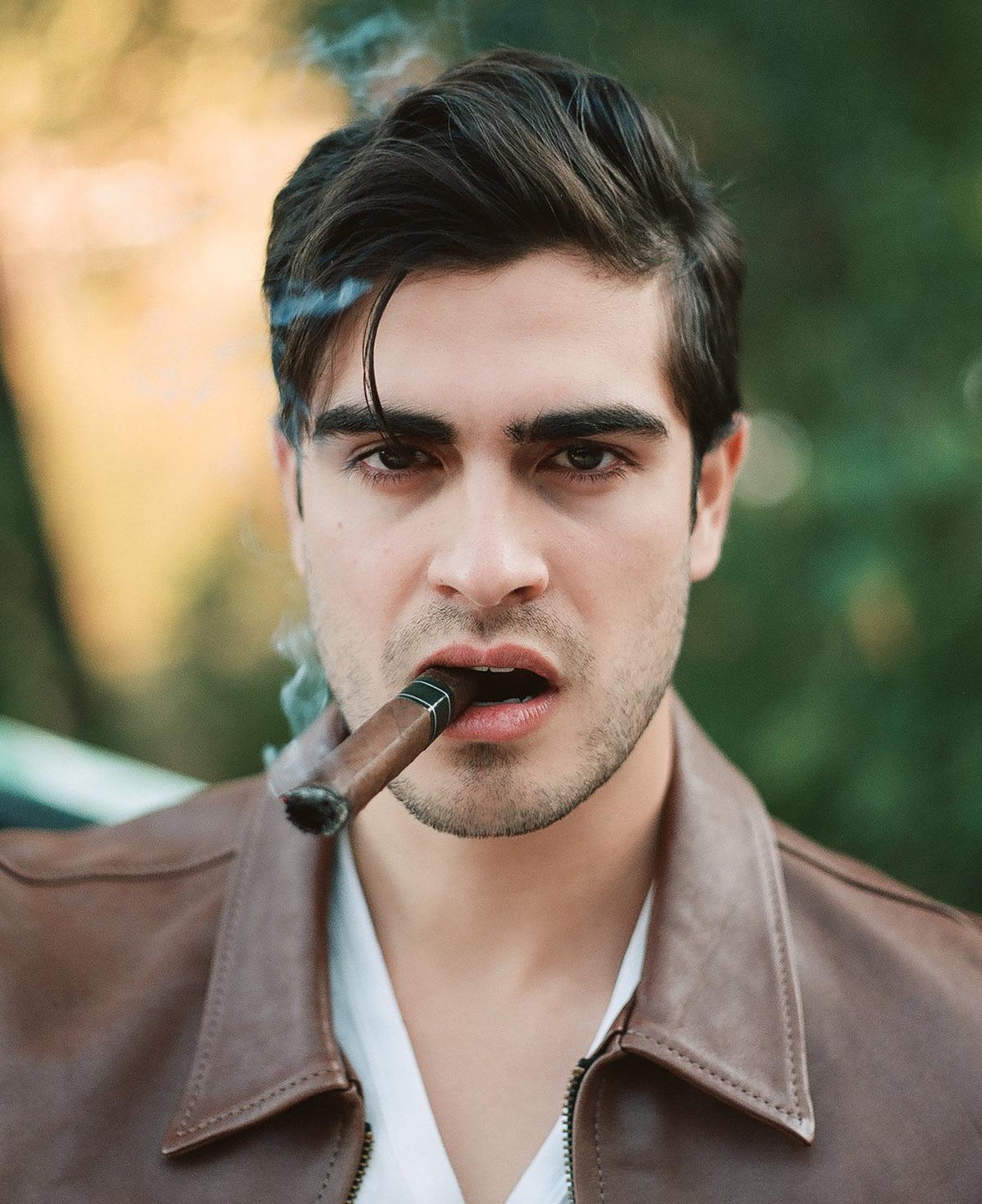 A man with dark hair and a brown leather jacket holds a lit cigar in his mouth, with smoke rising from it.