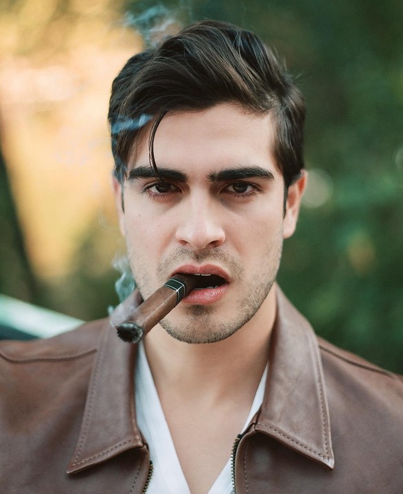 A man with dark hair and a brown leather jacket holds a lit cigar in his mouth, with smoke rising from it.