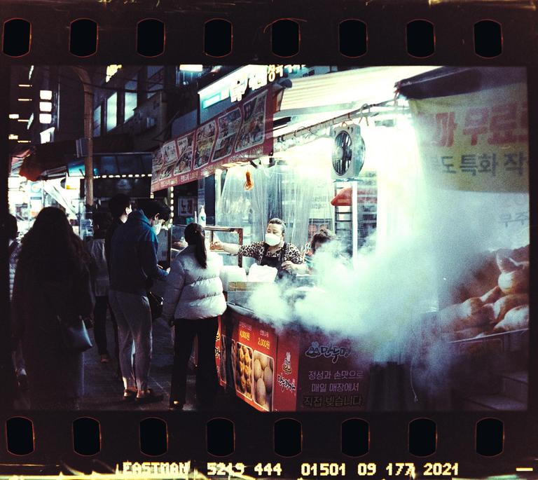 A woman wearing a face mask serves food from a steaming street stall at night.
