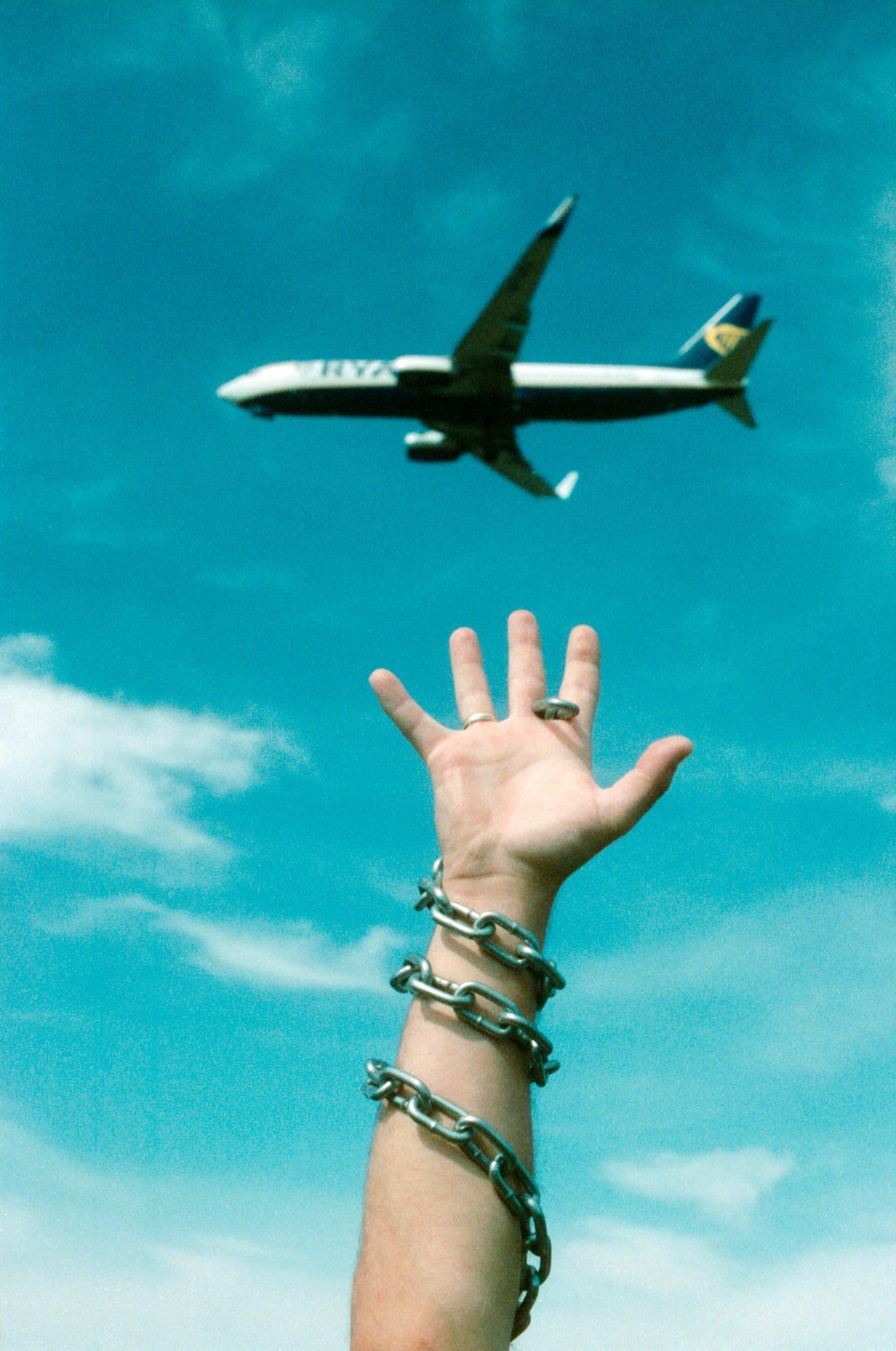 A person's arm wrapped in chains reaches towards an airplane flying in a bright blue sky.