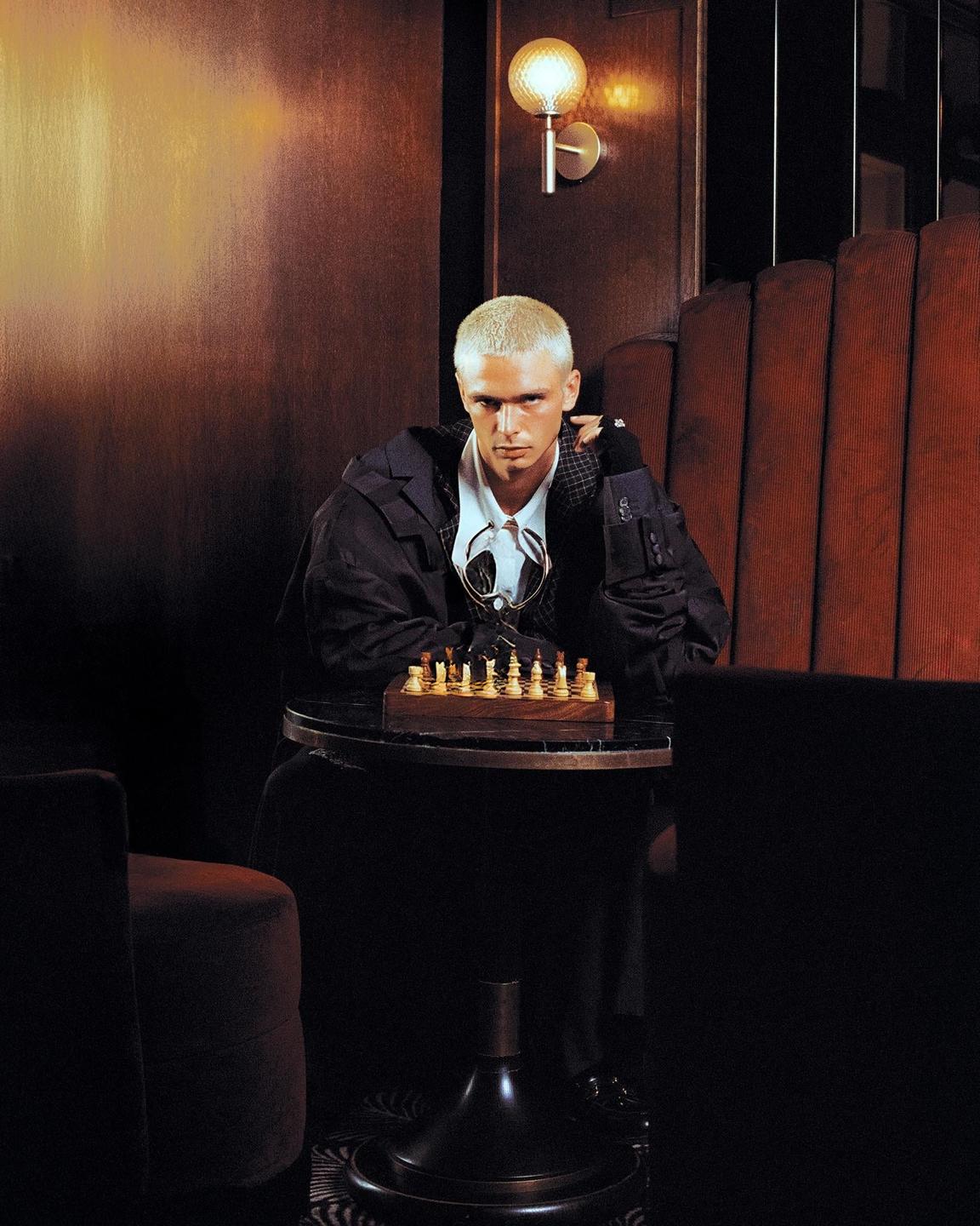 A man with bleached hair sits at a small table with a chessboard in a dimly lit room.