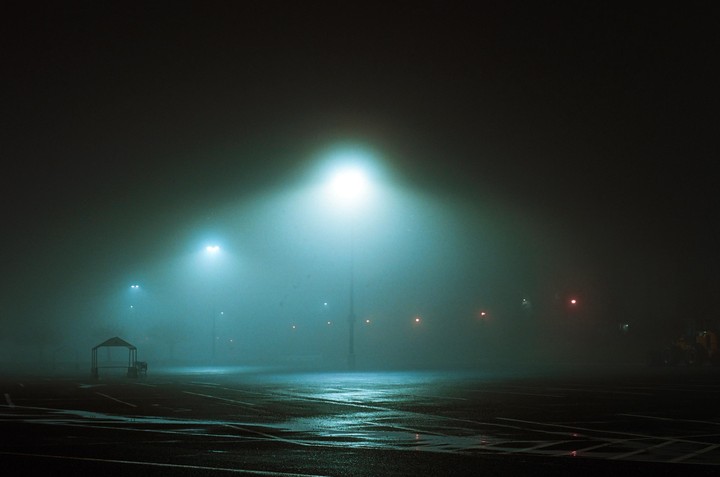 A foggy parking lot at night is illuminated by streetlights.