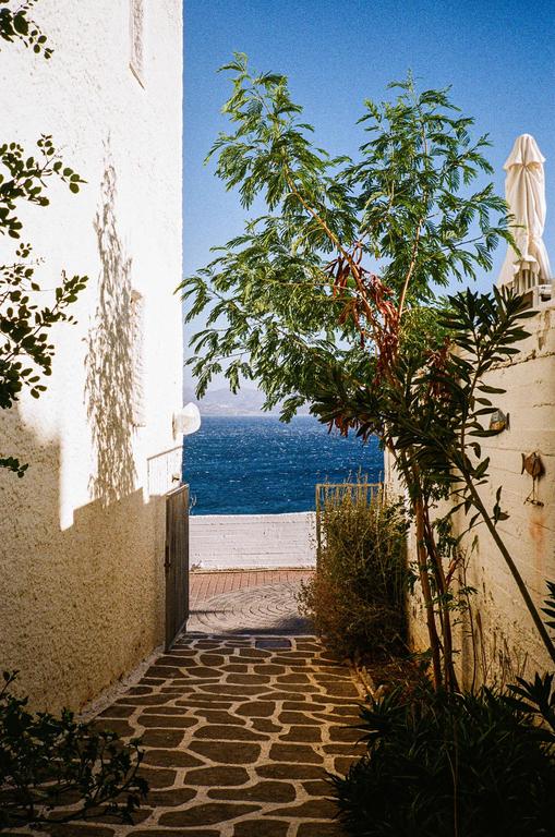 A narrow alleyway with white walls and a patterned stone path leads to a view of the ocean and distant mountains.