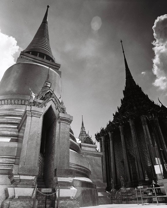 Two ornate temple spires rise towards a cloudy sky, with a large stupa on the left and a dark, intricate building on the right.