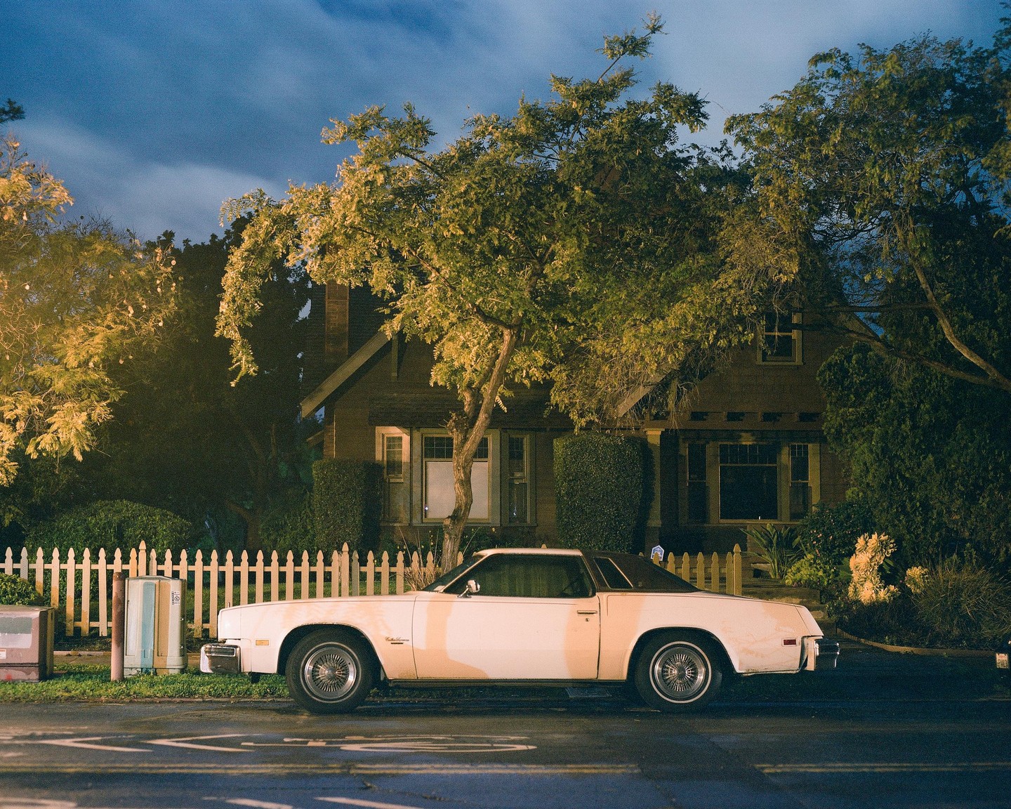 A white car is parked on the street in front of a house with a white picket fence at night.