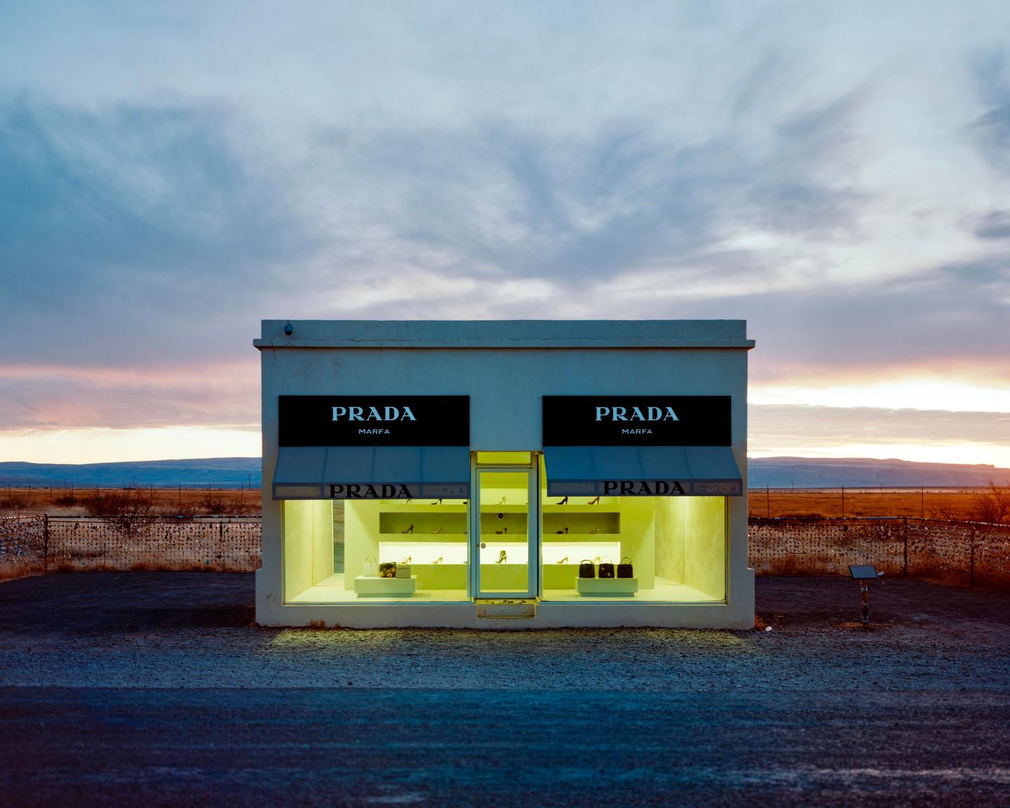 A small building with "PRADA MARFA" signs on its facade stands in a desolate landscape under a colorful sky.