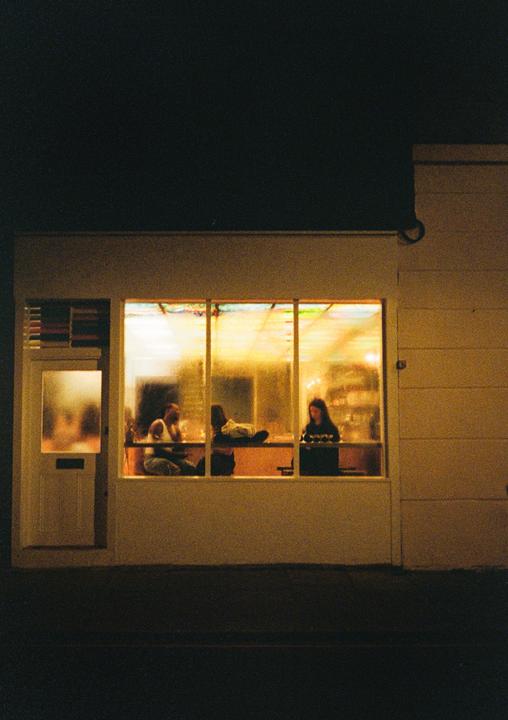 Three people are visible through a brightly lit window of a building at night.