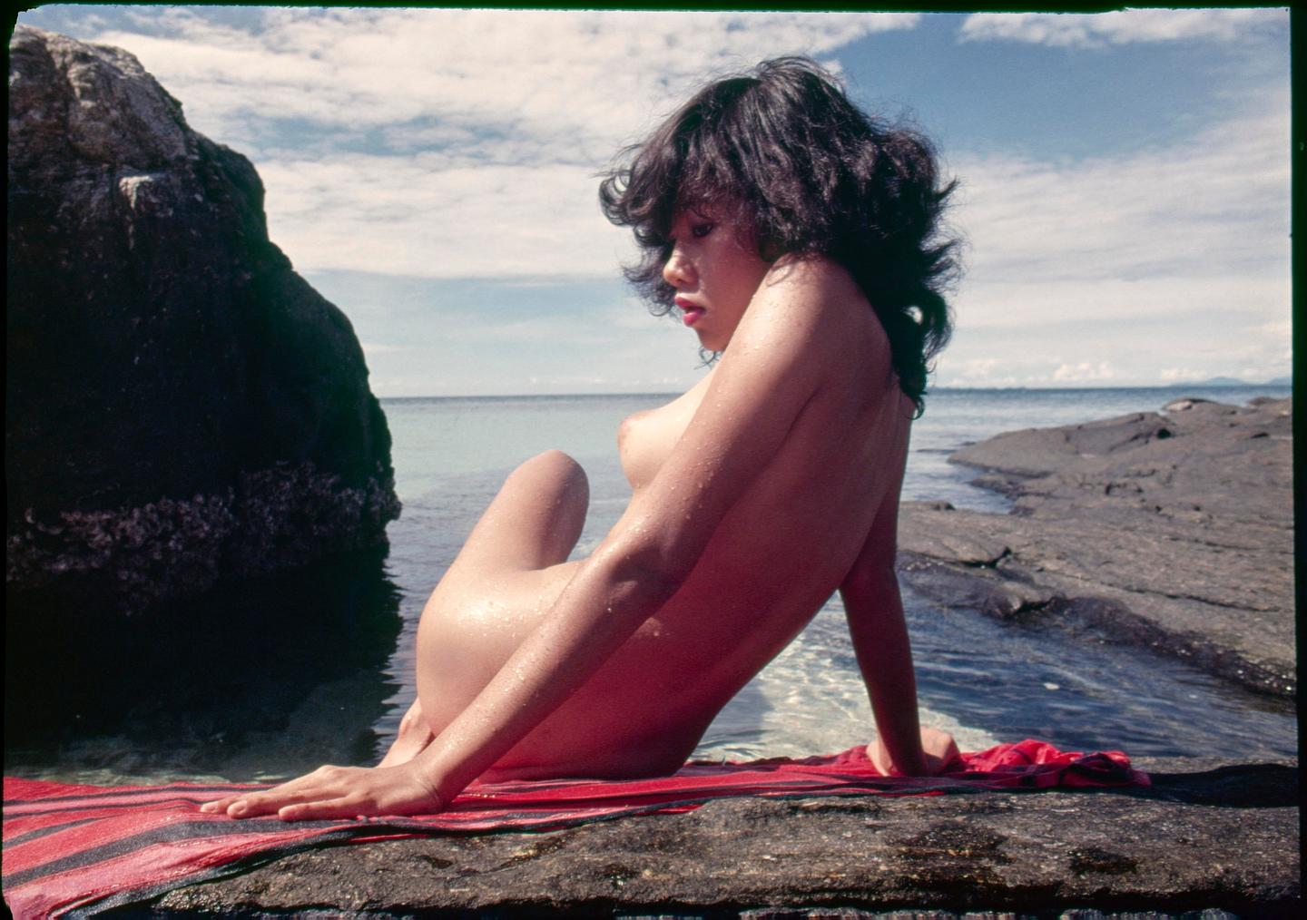 A woman sits nude on a red and black striped towel on a rocky shore with the ocean and a cloudy sky in the background.