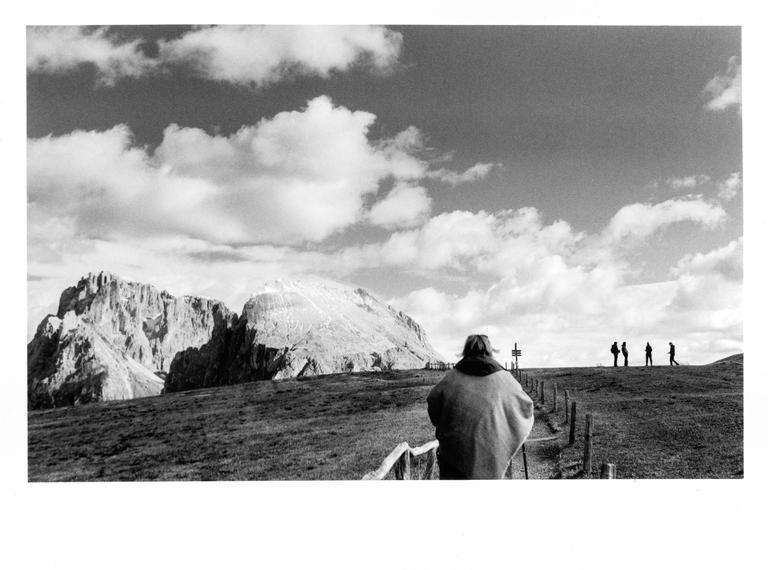 A person walks along a path with a mountain range in the background and other people in the distance.