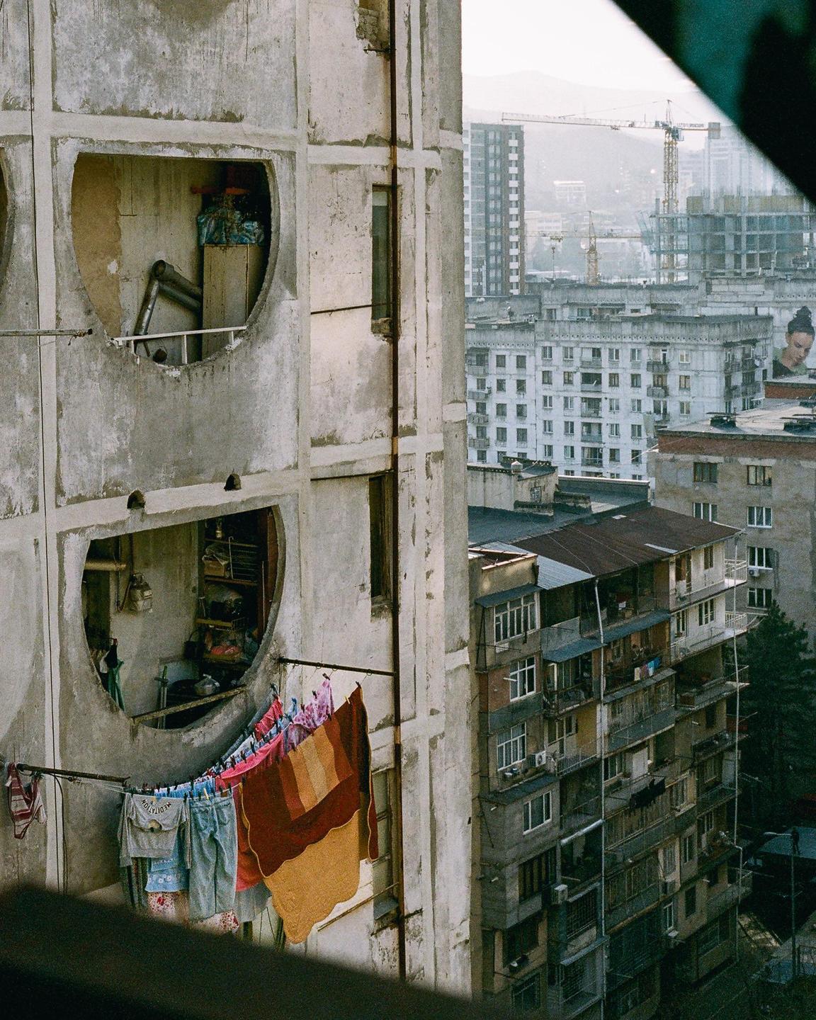 Laundry hangs on a clothesline outside a concrete building with circular windows overlooking a city.