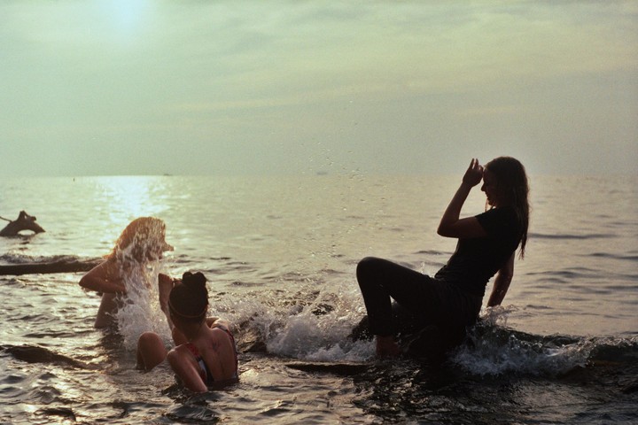 Three women are playing in the water at sunset, with one woman splashing water and another shielding her face.
