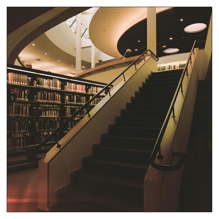 A wide staircase with a dark carpet and a metal handrail leads up to a landing in a library with bookshelves and a curved ceiling.