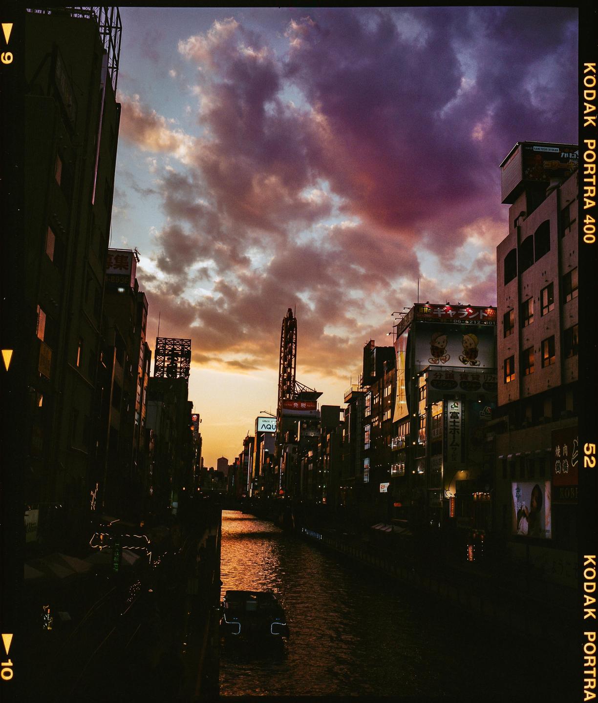 A river flows through a city with tall buildings and illuminated signs under a dramatic sunset sky.