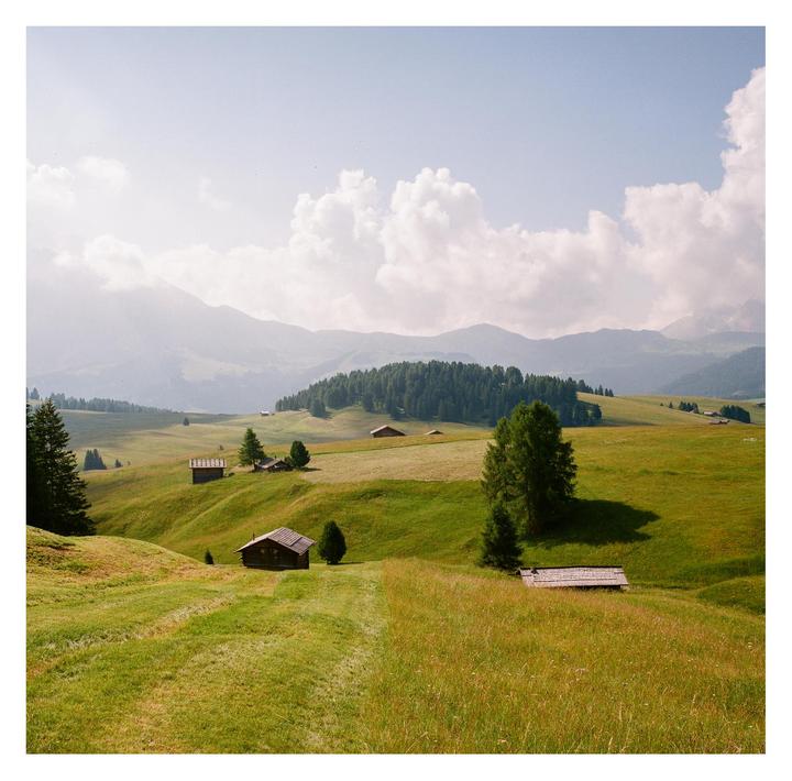 Small wooden huts are scattered across a green, hilly landscape with mountains and a forest in the background under a cloudy sky.