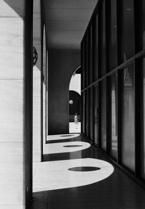 A person walks through a sunlit hallway with arches and shadows.