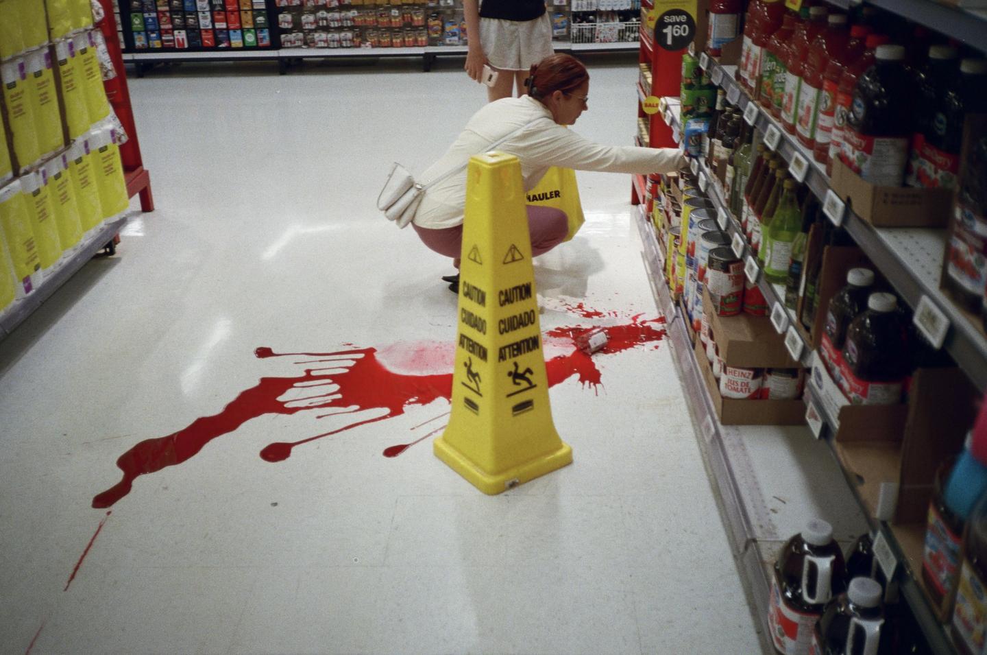 A woman crouches in a supermarket aisle next to a yellow caution cone and a large red spill on the floor.