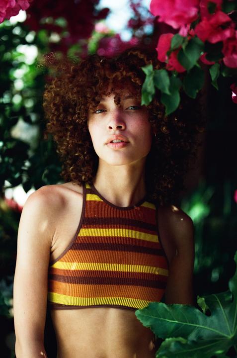 A woman with curly hair and a striped top is surrounded by pink flowers and green foliage.