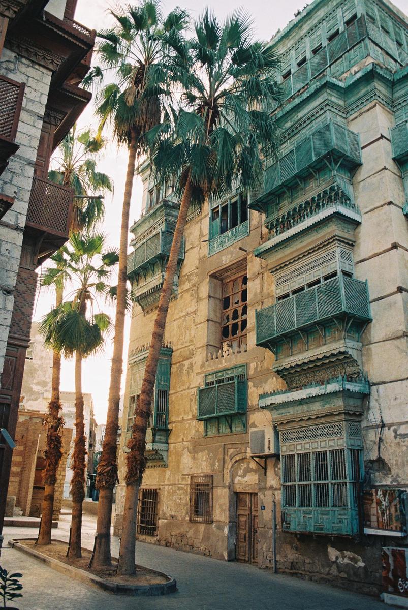 Palm trees line a narrow street with old buildings featuring ornate wooden balconies and shutters.