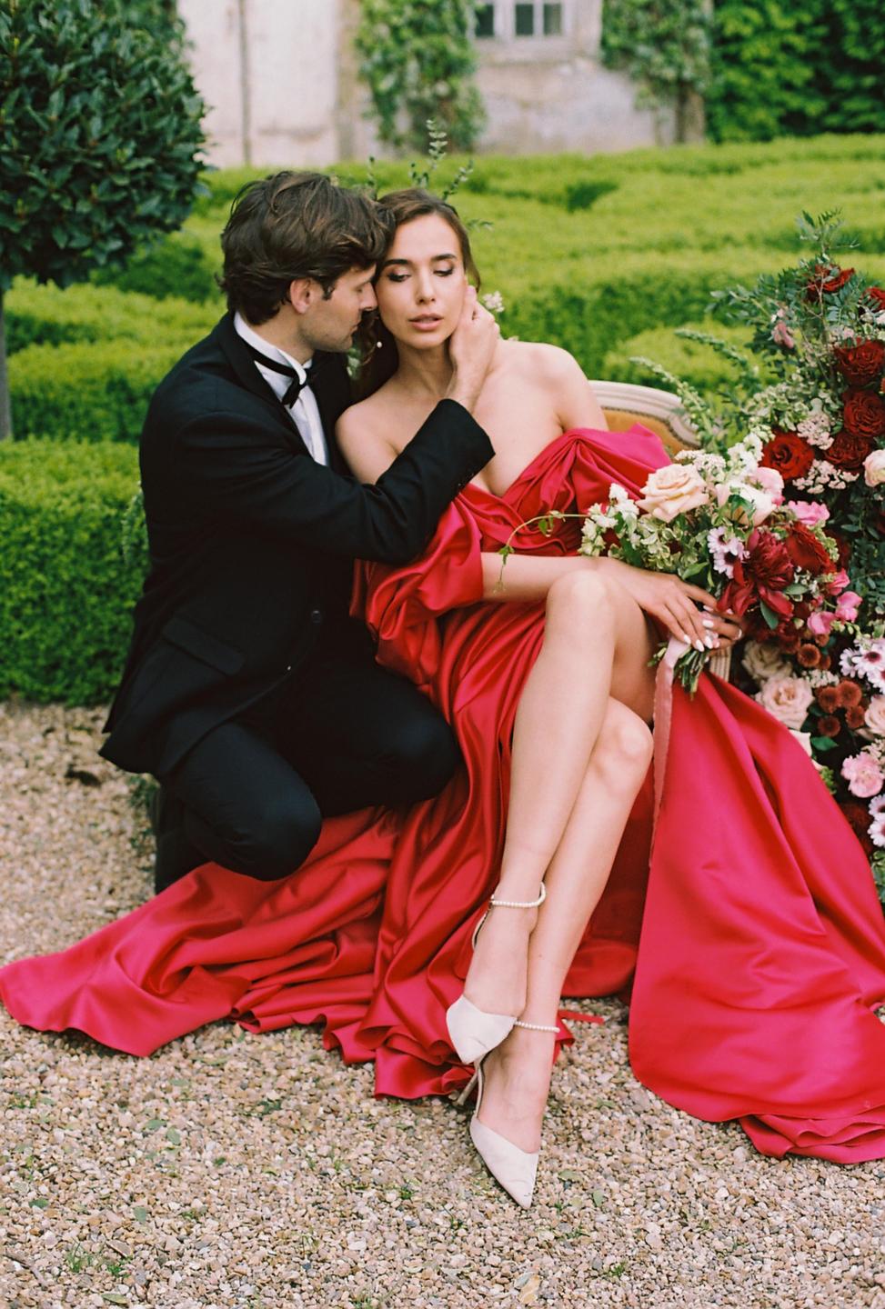 A man in a tuxedo kneels beside a woman in a red dress, who is seated on a couch with a bouquet of flowers next to her.