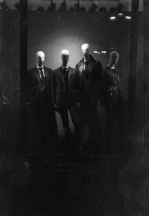 Four mannequins in suits are displayed in a dark storefront window.