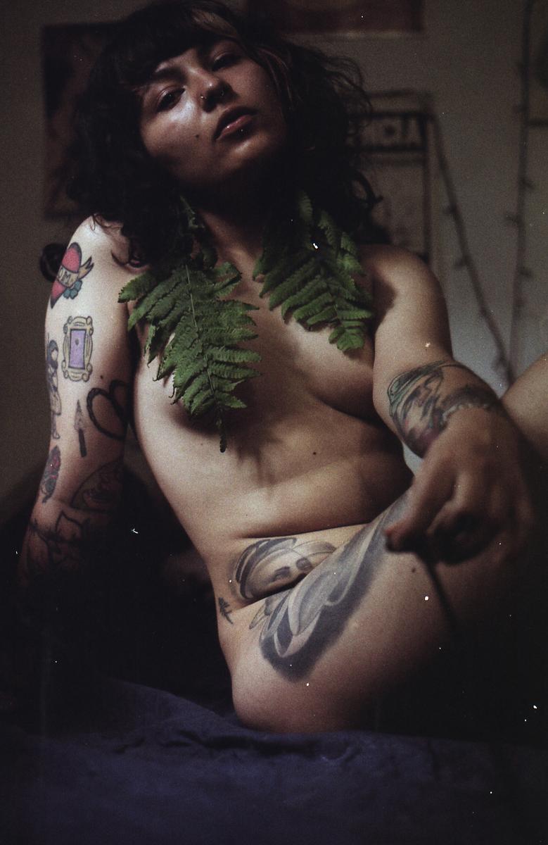 A woman with tattoos and fern leaves around her neck is reclining on a dark surface.
