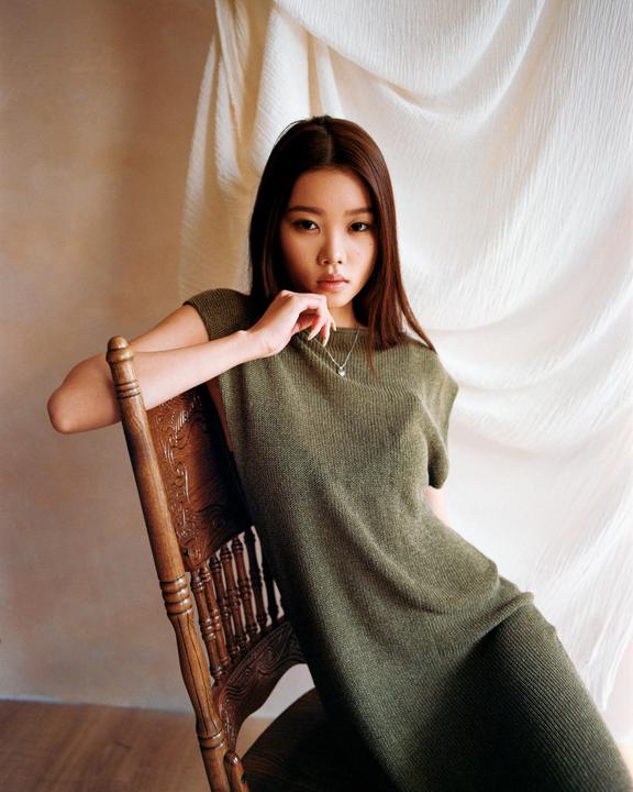 A woman in a green dress sits on a wooden chair with her arm resting on the back of the chair.