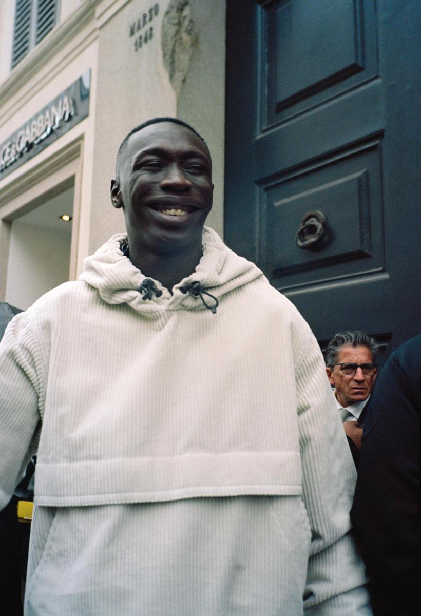 A man in a cream corduroy hoodie smiles at the camera on a street with a building and a dark door in the background.