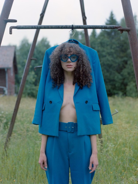 A woman with curly hair and blue sunglasses stands in a field with a blue jacket hanging on a metal frame above her.
